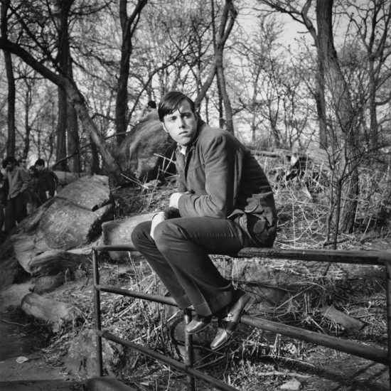 A black and white photograph of a man sitting on a metal railing in a park.