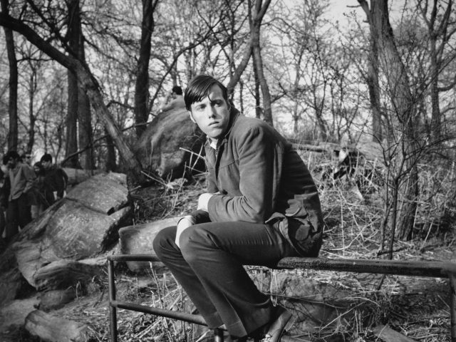 A black and white photograph of a man sitting on a metal railing in a park.