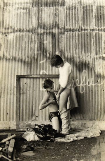 A black and white photograph of a man receiving oral sex in a post industrial setting.