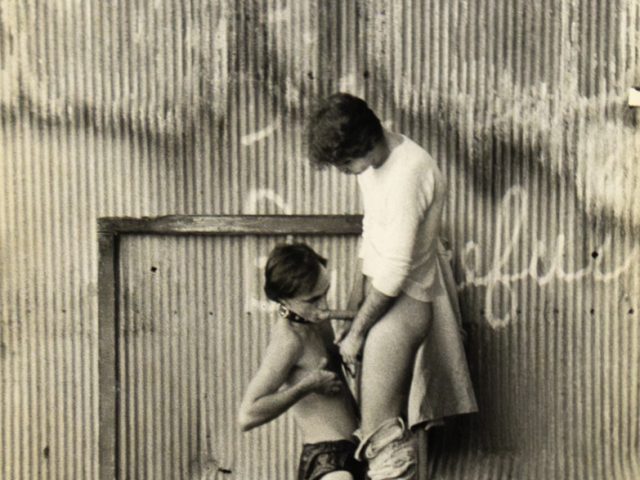 A black and white photograph of a man receiving oral sex in a post industrial setting.