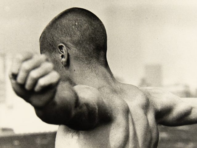 A black and white photograph of a boy with a shaved head with arms outstretched standing in front of a blurred skyline.