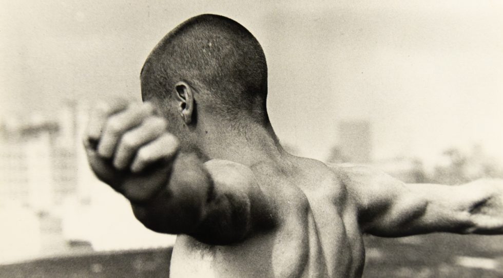 A black and white photograph of a boy with a shaved head with arms outstretched standing in front of a blurred skyline.