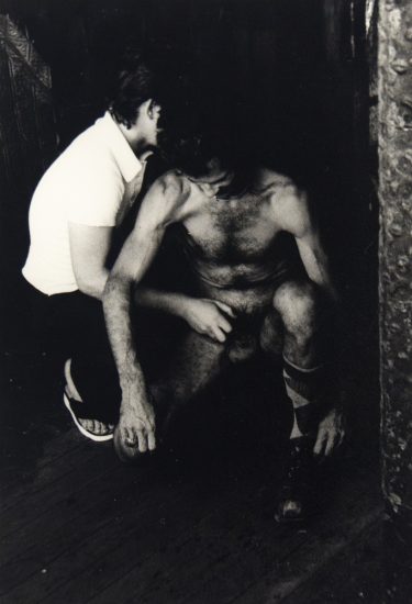 A black and white photograph of a nude man being fondles by a man in a white shirt.