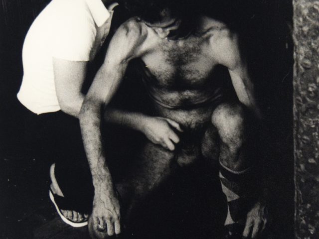 A black and white photograph of a nude man being fondles by a man in a white shirt.