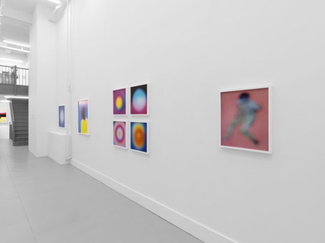 A photograph of an installation of colorful abstract works on a white gallery wall.