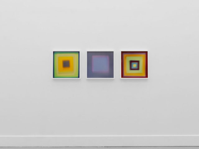 A photograph of an installation of colorful abstract works on a white gallery wall.