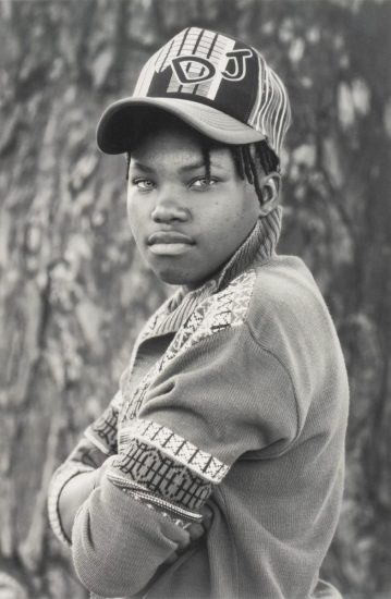 A black and white portrait of a young person in a cap with their arms crossed.