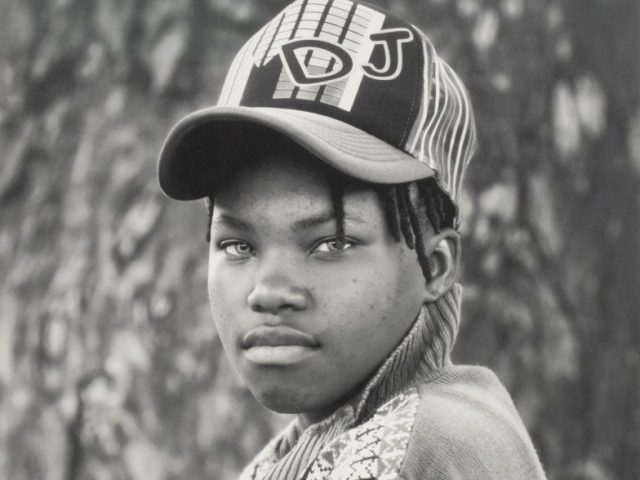 A black and white portrait of a young person in a cap with their arms crossed.