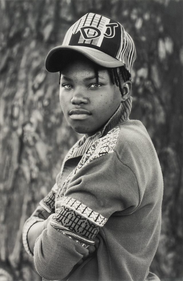 A black and white portrait of a young person in a cap with their arms crossed.