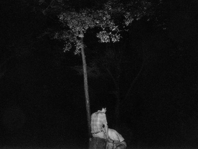 A black and white photograph of two men by a tree in the darkness.