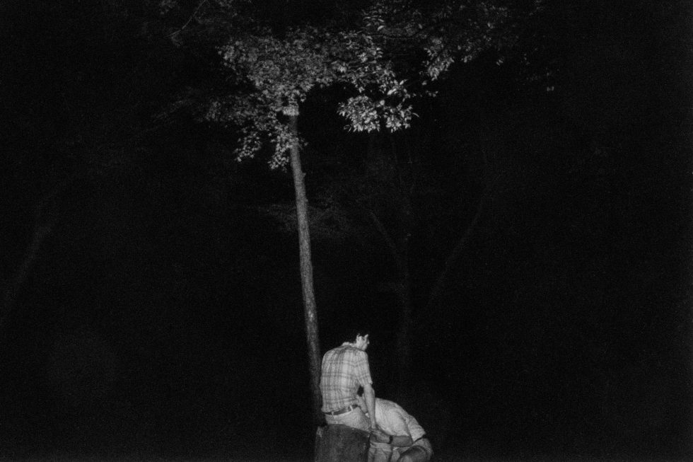 A black and white photograph of two men by a tree in the darkness.