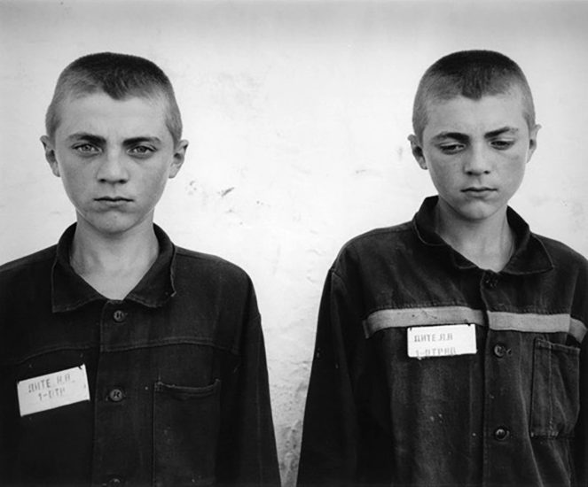 A black and white portrait of two boys in prison uniforms in front of a white wall.