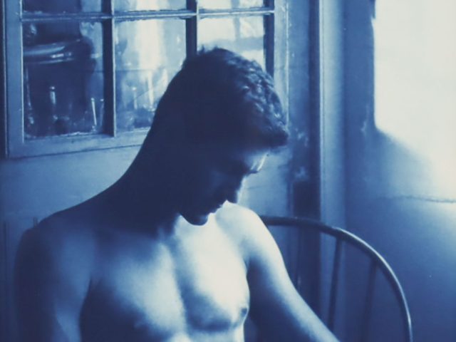 A cyanotype photograph of a shirtless man sitting in a chair by a window bathed in natural light.