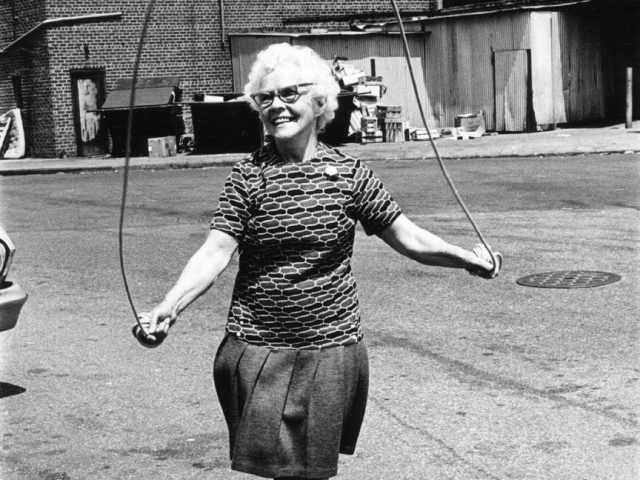 A black-and-white photograph of an older woman jumping rope on the street.