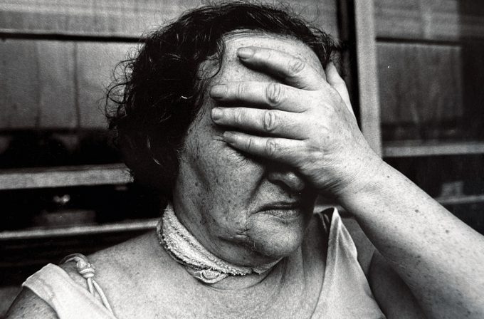A black and white photograph of an older woman covering her face with her hand.