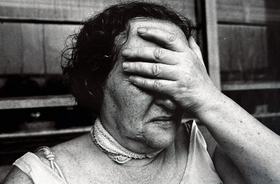 A black and white photograph of an older woman covering her face with her hand.