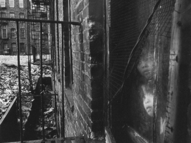 A black and white photograph of two children peering out a window onto a fire escape.