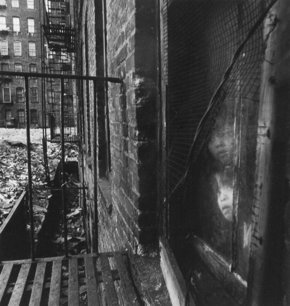 A black and white photograph of two children peering out a window onto a fire escape.