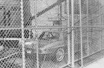 A black and white photograph of a car behind a chain link fence.