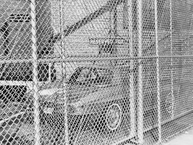 A black and white photograph of a car behind a chain link fence.