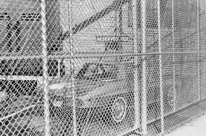 A black and white photograph of a car behind a chain link fence.