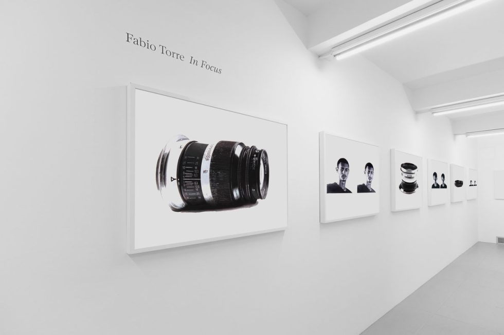 A photograph of a white-walled art gallery with black and white paintings with white frames on view.