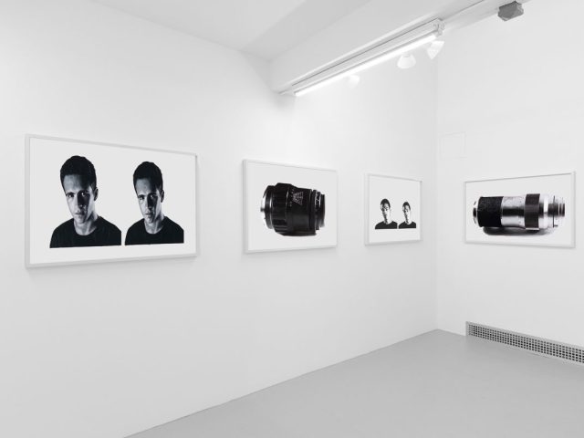 A photograph of a white-walled art gallery with black and white paintings with white frames on view.