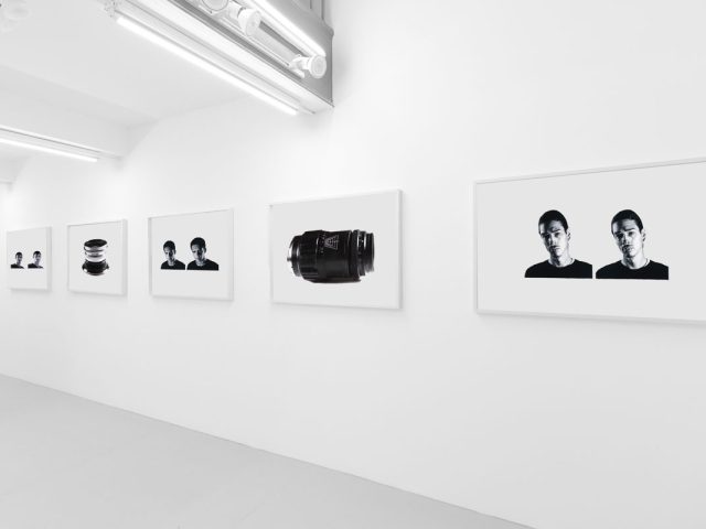A photograph of a white-walled art gallery with black and white paintings with white frames on view.