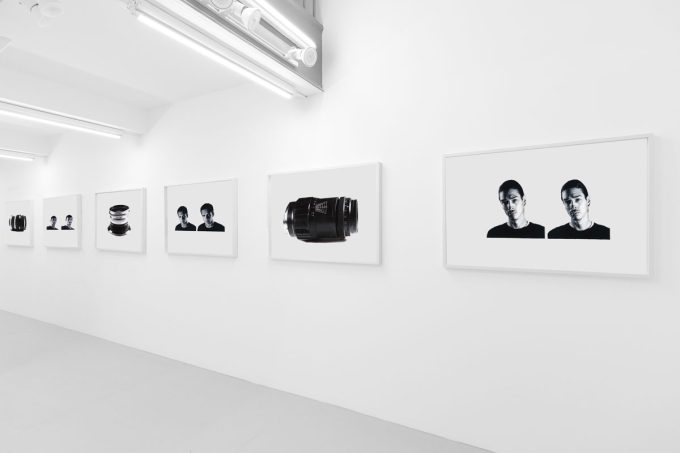 A photograph of a white-walled art gallery with black and white paintings with white frames on view.