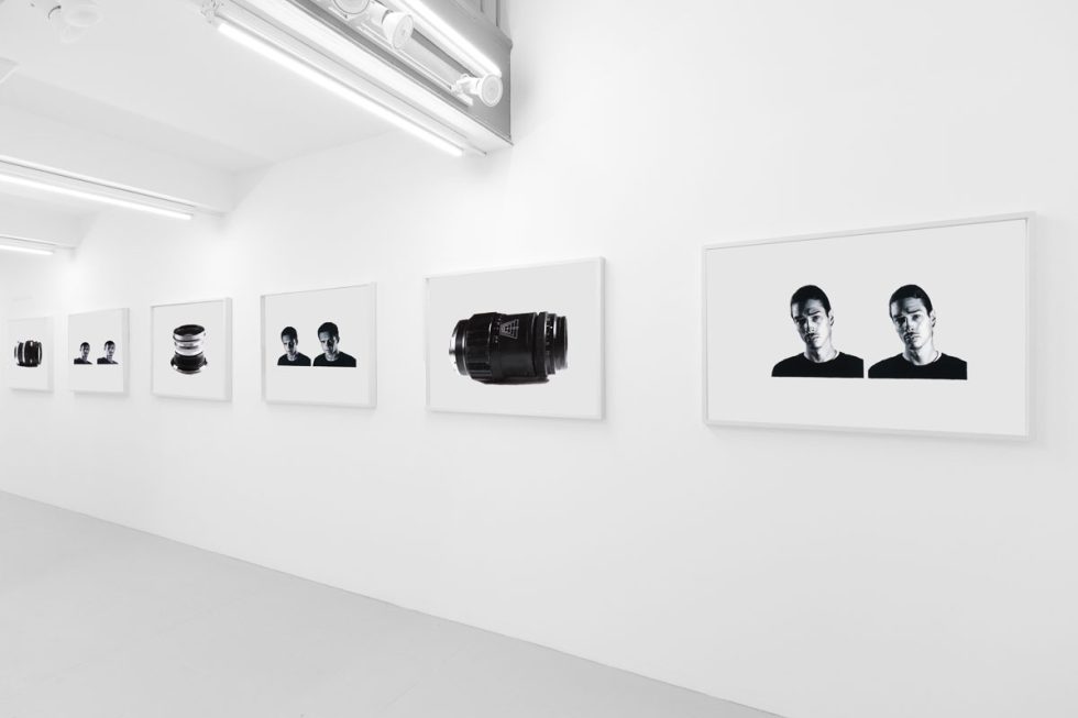 A photograph of a white-walled art gallery with black and white paintings with white frames on view.