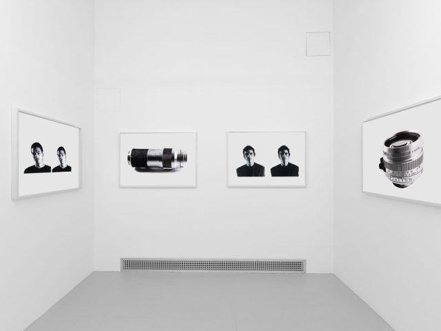 A photograph of a white-walled art gallery with black and white paintings with white frames on view.