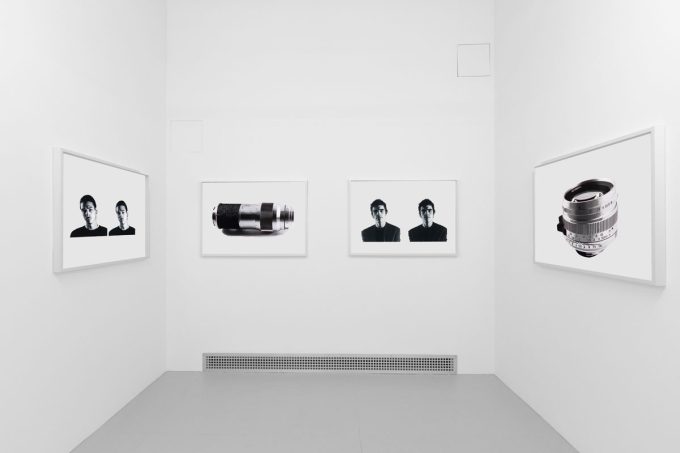 A photograph of a white-walled art gallery with black and white paintings with white frames on view.