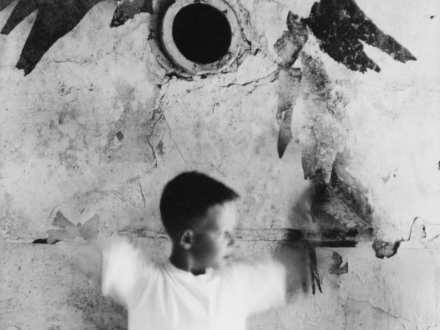 A black and white photograph of a young boy in motion against a distressed wall.