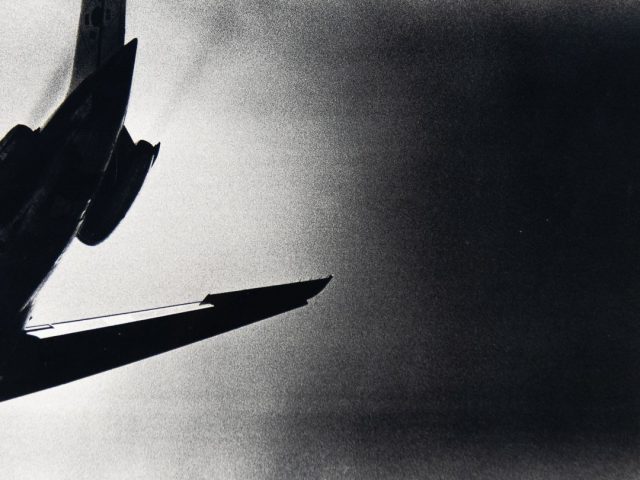 A black and white photograph of the tail of a plane seen from below.