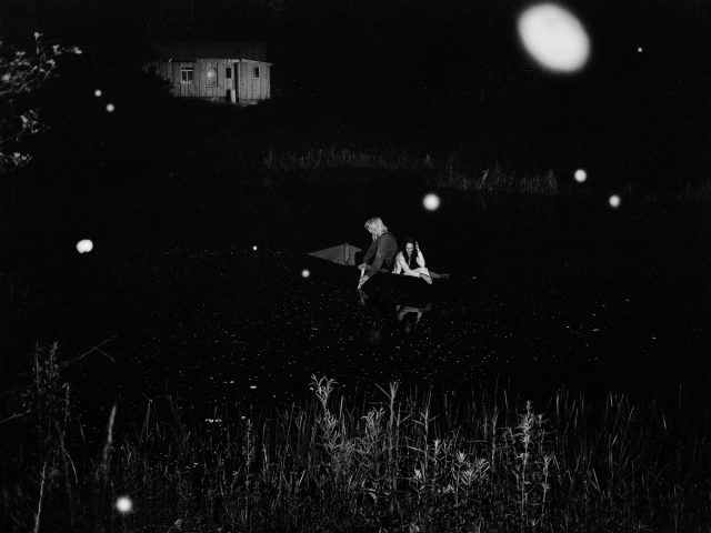 A black and white photograph of two people in a boat on a pond at night.