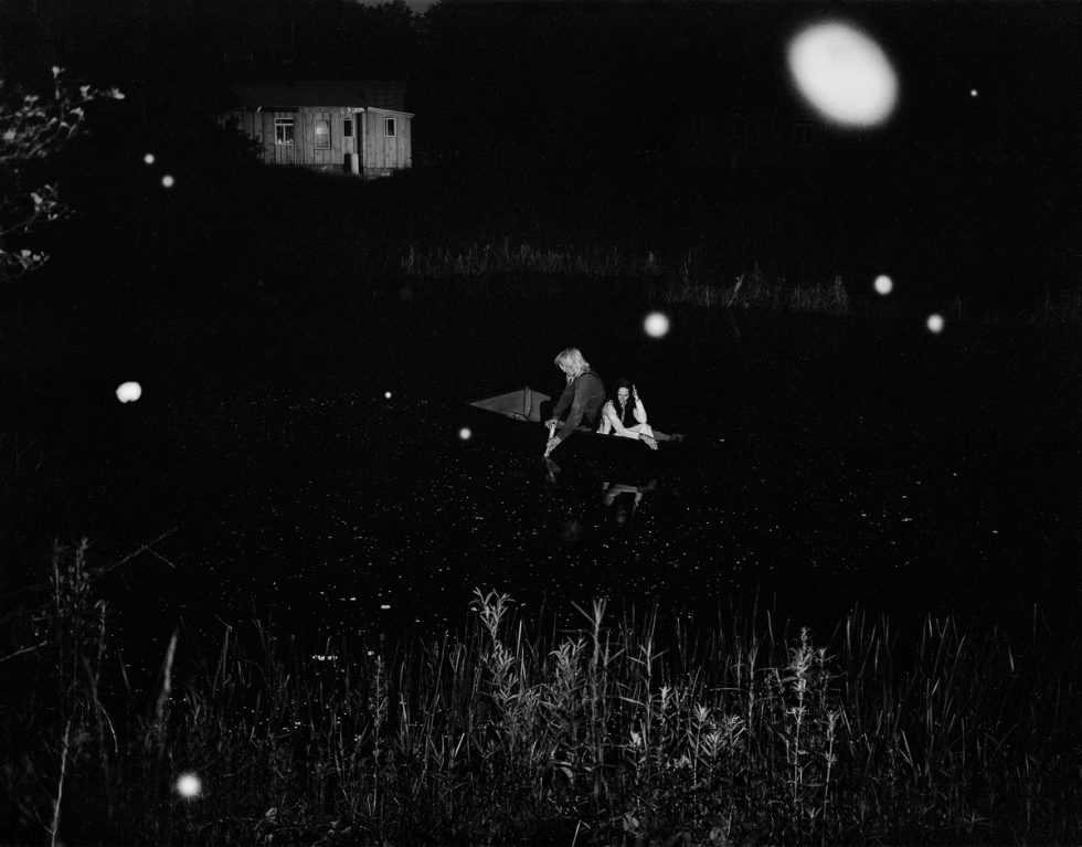 A black and white photograph of two people in a boat on a pond at night.