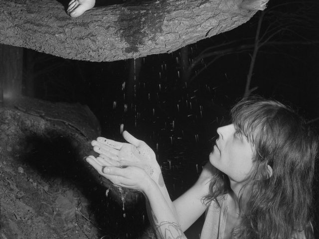 A black and white photograph of a nude woman in a tree urinating on the hands of another woman below her.