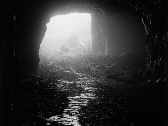 A black and white photograph looking out of a dark cave with light streaming in from outside.