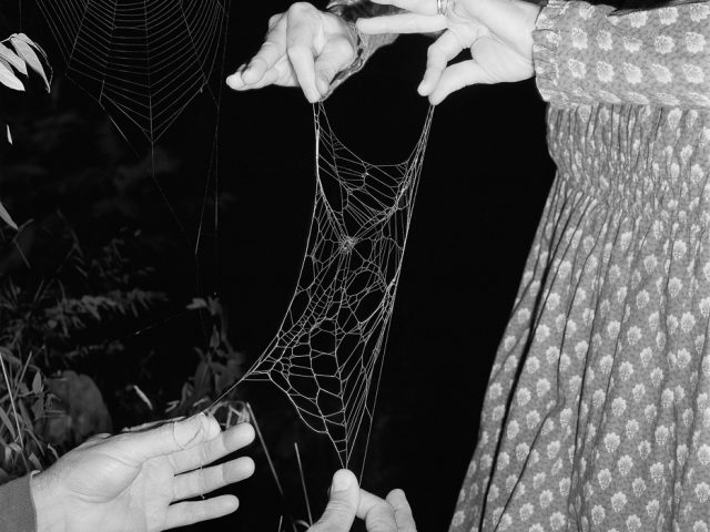 A black and white photograph of a woman in a patterned dress holding the edges of a spider web.