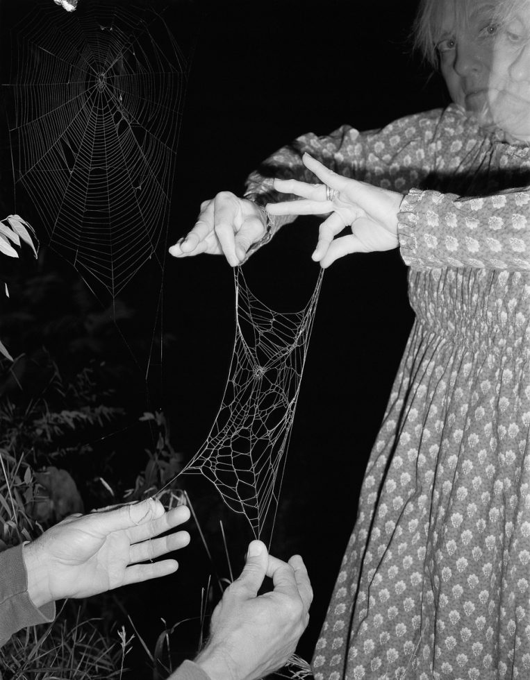 A black and white photograph of a woman in a patterned dress holding the edges of a spider web.