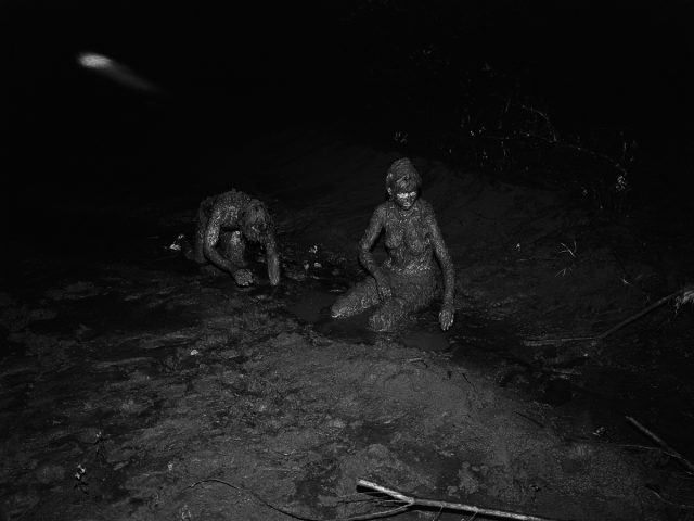 A black and white photograph of two nude figures completely covered in mud crawling on the ground.