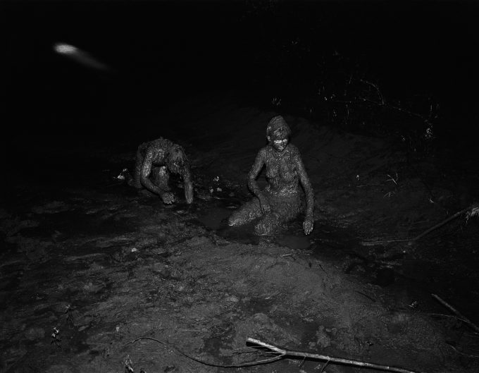 A black and white photograph of two nude figures completely covered in mud crawling on the ground.