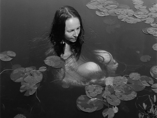 A black and white photograph of a woman sitting in a pond surrounded by lily pads.