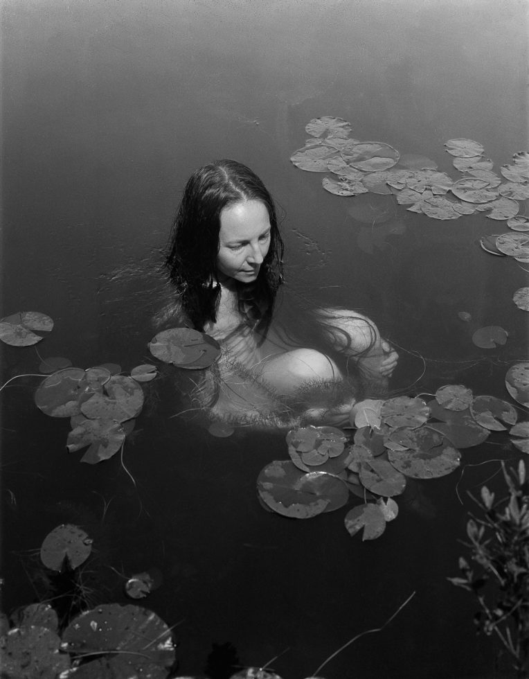 A black and white photograph of a woman sitting in a pond surrounded by lily pads.