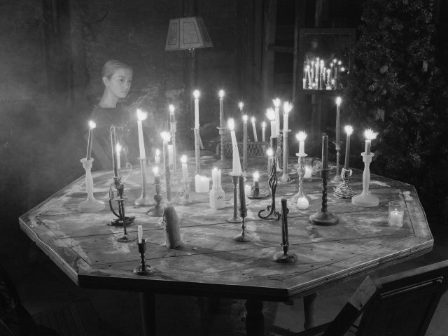 A black and white photograph of a woman sitting by a table covered in burning candles.