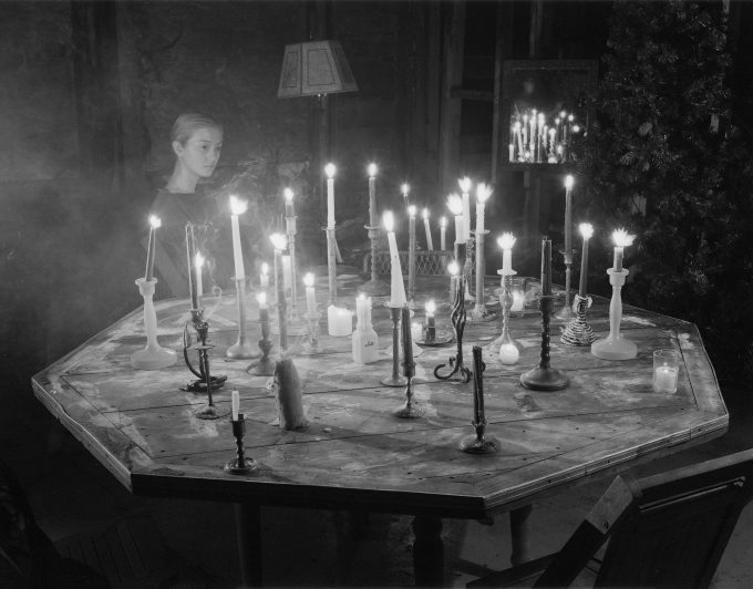 A black and white photograph of a woman sitting by a table covered in burning candles.