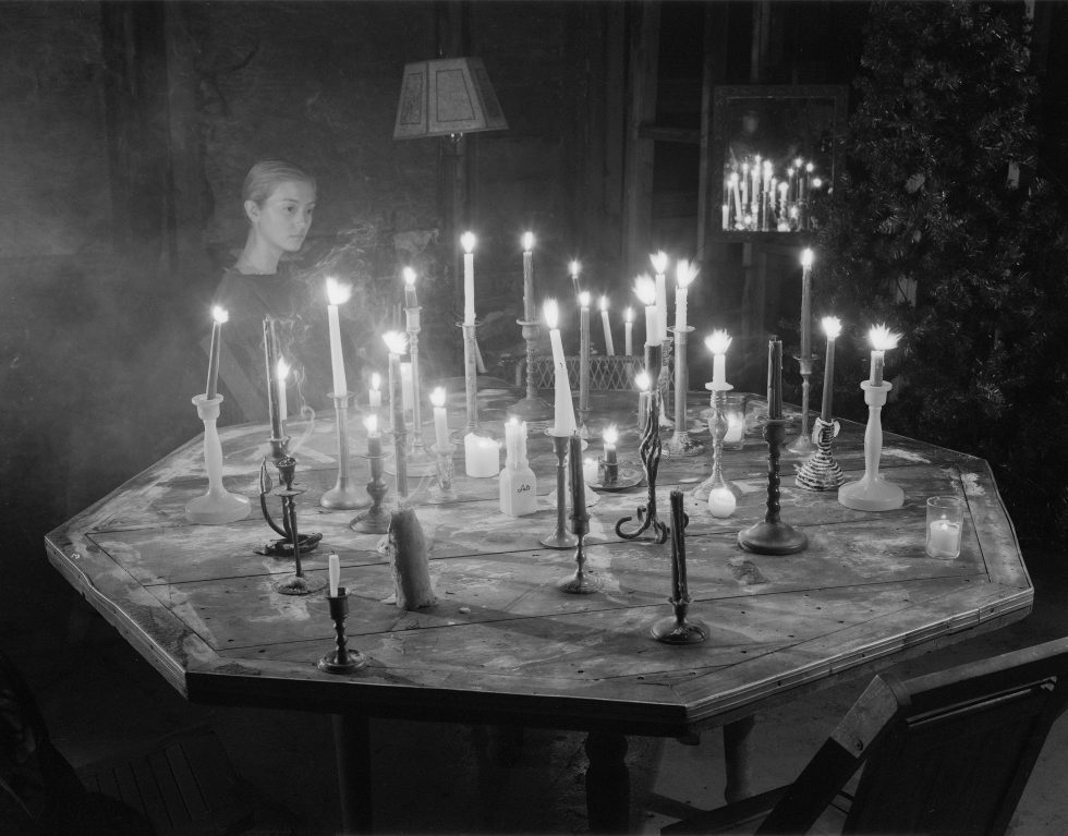 A black and white photograph of a woman sitting by a table covered in burning candles.