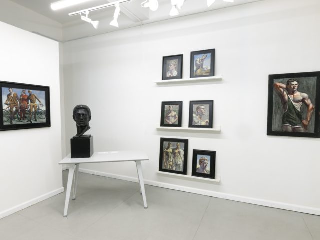 This image shows the walls of a gallery where eight framed paintings hang. On a table, a black statue of a man's head is displayed.