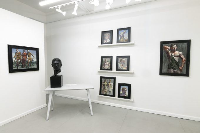 This image shows the walls of a gallery where eight framed paintings hang. On a table, a black statue of a man's head is displayed.