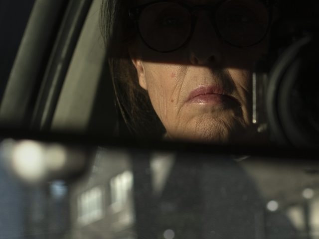A photograph of a woman driving seen through the rearview mirror of a car.